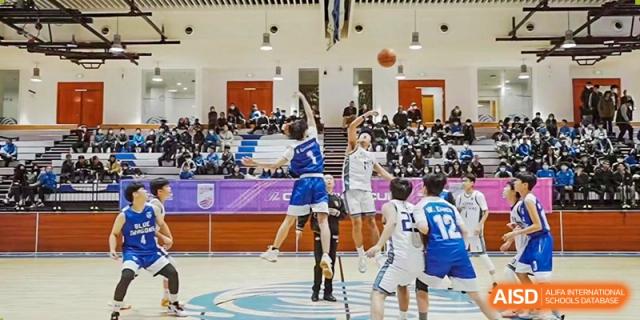 A player in a white and blue jersey for Xi'an Liangjiatan International School jumps high to shoot a basketball during a game in a packed gymnasium, with other players and spectators visible.
