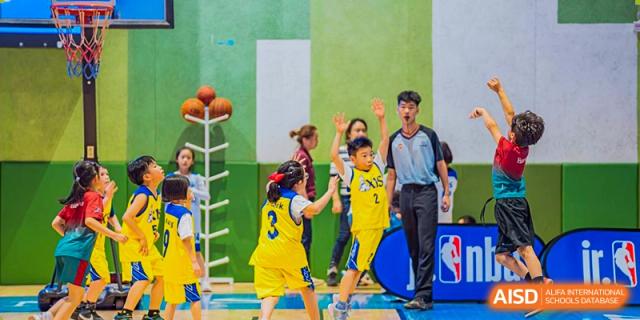 Players from Xi'an Liangjiatan International School, wearing yellow and blue uniforms, watch as a referee and another player stand on the court during a stoppage or play in a basketball game.
