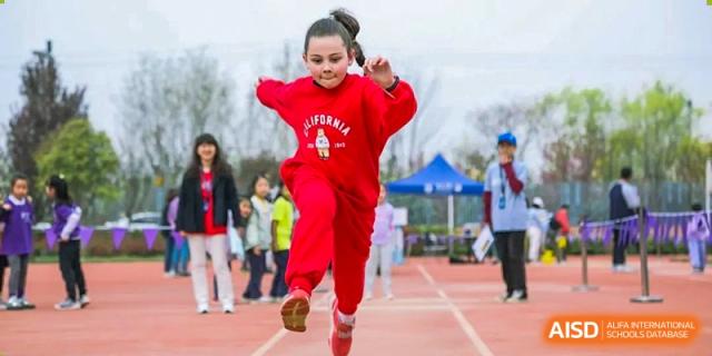A girl wearing a red sweatshirt for Xi'an Liangjiatan International School runs and jumps on a red outdoor track during an athletic event.