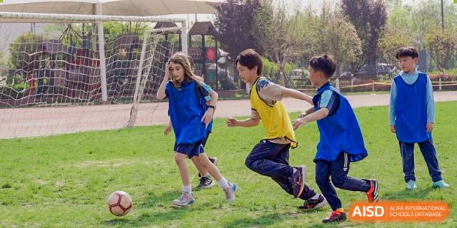 Two young students from Xi'an Liangjiatan International School, one in blue and one in yellow, running and smiling on a grassy field during an outdoor activity.