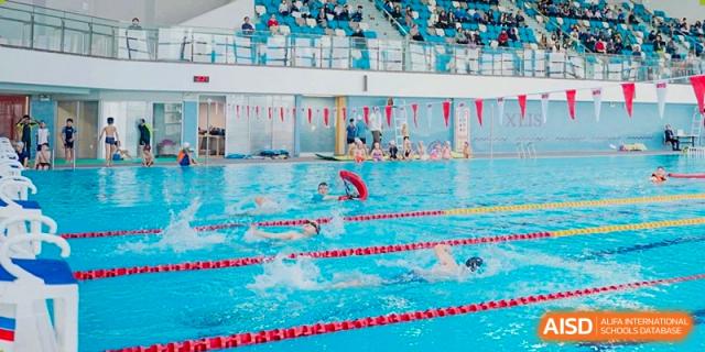 A student from Xi'an Liangjiatan International School competes in a formal swimming race in a pool with lane lines, with spectators visible in the stands.