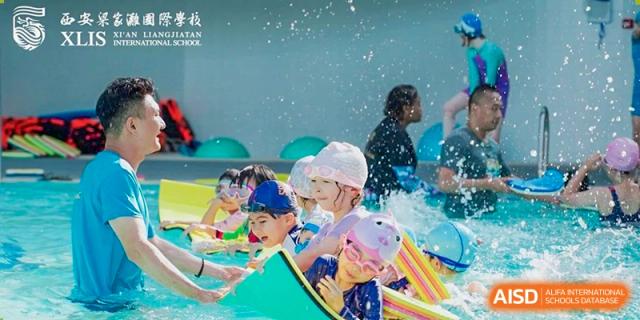 Young children from Xi'an Liangjiatan International School wearing swim caps and goggles splashing happily in a pool during a lesson.