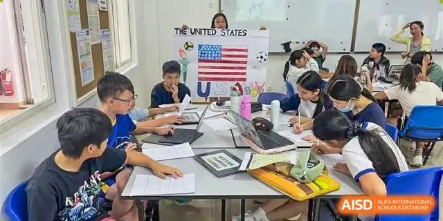 Xiamen International School students working together on laptops and papers in a classroom, with a US flag visible.