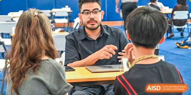 A male teacher or staff member with glasses at Xiamen International School talking with a group of students at a table.
