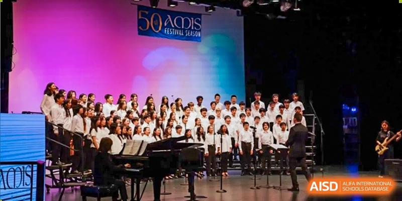 The Xiamen International School choir performing on stage, with a piano visible in the foreground.