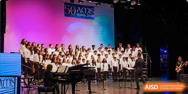 The Xiamen International School choir performing on stage, with a piano visible in the foreground.