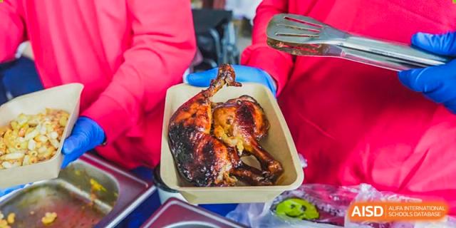 A whole roasted chicken being held with tongs in a red tray at a Xiamen International School food event.