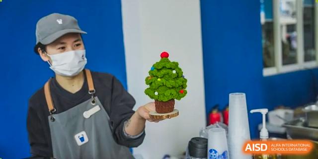 A hand holding a small, decorative Christmas tree made of leaves or greenery at a Xiamen International School cultural event.