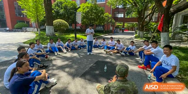 A teacher or instructor speaking to a circle of Yali High School International Department students and an officer seated on the ground outdoors.