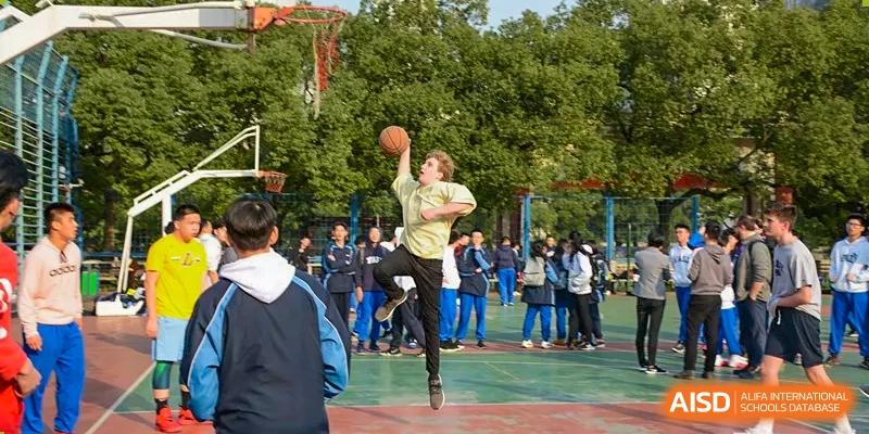 A male student in a light green shirt jumps high to shoot a basketball on an outdoor court surrounded by spectators at Yali High School International Department.