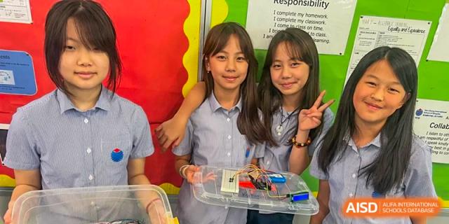 Two female students at Yew Chung International School of Qingdao smiling while holding a tray of electronics or science components in the classroom.