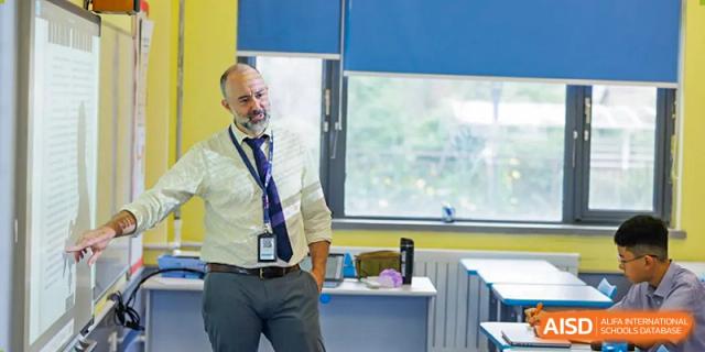A male teacher at Yew Chung International School of Qingdao with a beard, wearing a white shirt and tie, standing casually in a classroom setting.
