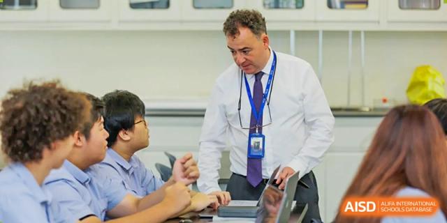 A male teacher at Yew Chung International School of Qingdao assisting a student with work on a digital tablet at a desk.