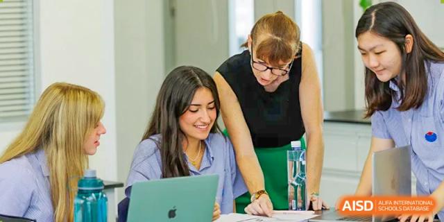 A female teacher at Yew Chung International School of Qingdao leaning over to help a female student with work on a laptop in the classroom.