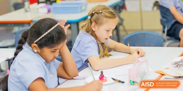 A young female student at Yew Chung International School of Qingdao in school uniform, concentrating on writing or an assignment at a desk.