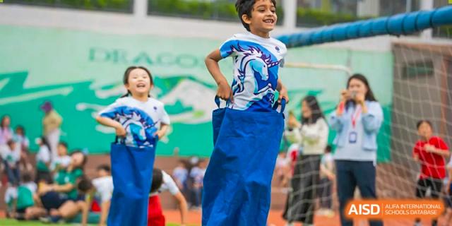 A young boy at Yew Chung International School of Qingdao participating enthusiastically in a sack race outdoors, wearing a blue sack and smiling.