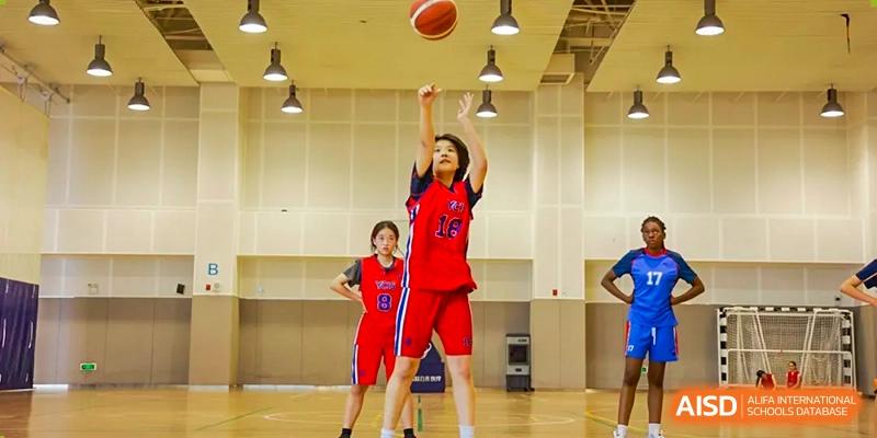 A student at Yew Chung International School of Qingdao in a red uniform shooting a basketball during an indoor game.