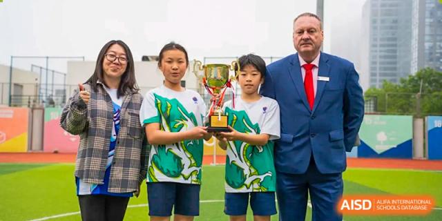 Two students at Yew Chung International School of Qingdao proudly holding up a trophy, standing next to a staff member in a suit.