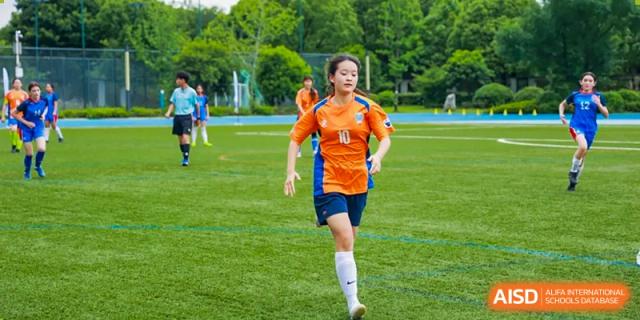 A female student at Yew Chung International School of Qingdao running on an outdoor sports field in an orange and blue uniform during a game.