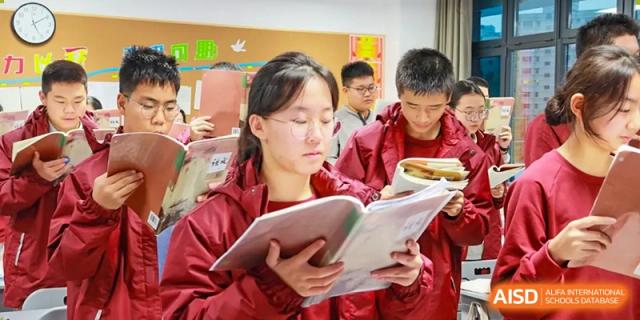 A female student focused on reading an open book in a classroom, surrounded by other students in red uniforms at Zibo MacDuffie Bilingual High School.