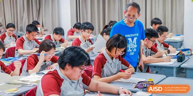 A classroom of students in red uniforms working on an assignment at their desks, with a teacher assisting them at Zibo MacDuffie Bilingual High School.