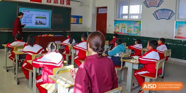 A classroom scene showing a teacher in a maroon uniform or sweater facing the class, with students working at their desks at Zibo MacDuffie Bilingual High School.
