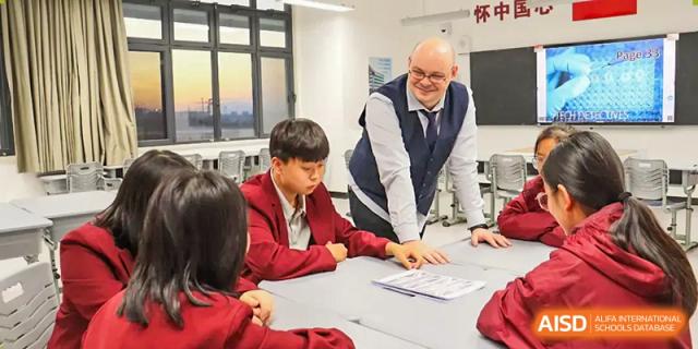 An international teacher, a Caucasian man, assisting a student in a red uniform with their work in a classroom at Zibo MacDuffie Bilingual High School.
