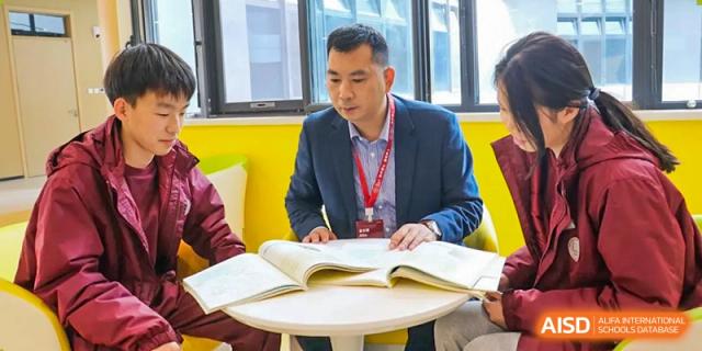A teacher or administrator, an Asian man, sitting at a table reviewing a large open textbook or document at Zibo MacDuffie Bilingual High School.