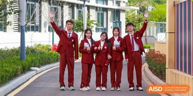 A line of five students in matching maroon school uniforms posing and smiling outdoors in front of a modern school building at Zibo MacDuffie Bilingual High School.