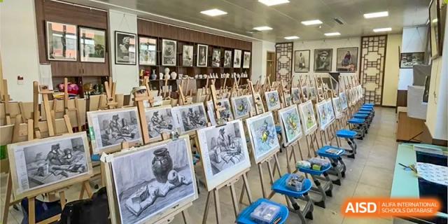 A brightly lit art room with numerous easels displaying various pencil and charcoal sketches, with framed artwork on the back wall at Zibo MacDuffie Bilingual High School.