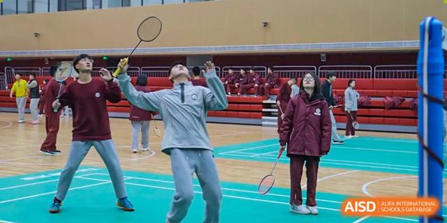 A male student in a gray tracksuit jumping to hit a shuttlecock or ball over a net during a sports activity indoors at Zibo MacDuffie Bilingual High School.