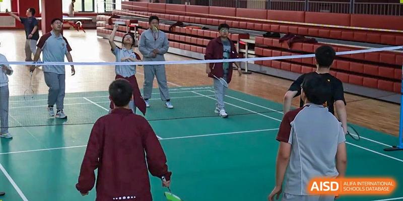 Two students playing a game of badminton on an indoor court, with others watching in the background at Zibo MacDuffie Bilingual High School.