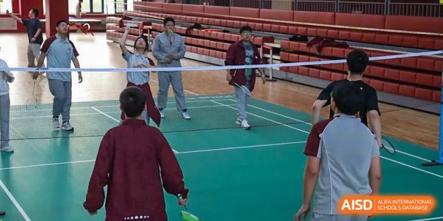 Two students playing a game of badminton on an indoor court, with others watching in the background at Zibo MacDuffie Bilingual High School.