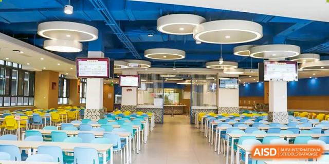 The brightly lit school cafeteria with rows of teal and yellow chairs and a blue ceiling, featuring modern circular pendant lights at Zibo MacDuffie Bilingual High School.
