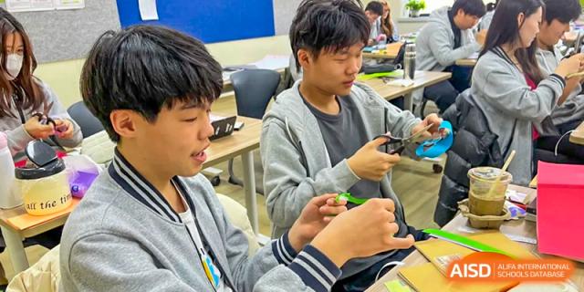 Two male students sitting side-by-side at a desk, focused on a hands-on project or activity using small objects, at Ulink College of Beijing.