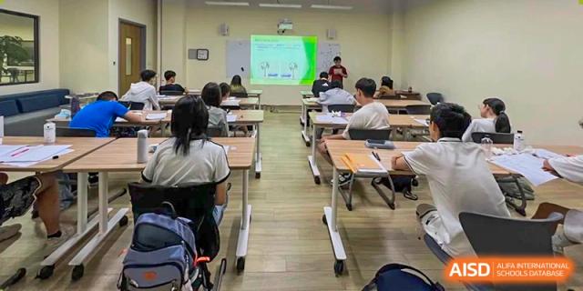 A teacher instructing students in a bright classroom, with a presentation visible on the whiteboard at the front of the room, at Ulink College of Beijing.