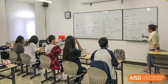 The backs of students seated at desks in a classroom, listening to a lesson with various mathematical equations written on the whiteboard, at Ulink College of Beijing.