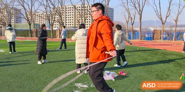 A male student in an orange puffer jacket swinging a rope or staff on an outdoor sports field, at Ulink College of Beijing.