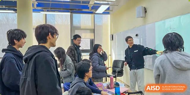 Students and teachers gathered around a desk in a classroom or common area, engaged in a focused discussion or consultation, at Ulink College of Beijing.