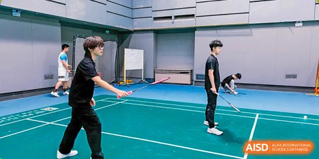 Two male students standing on an indoor blue court, preparing to play badminton with rackets, at Ulink College of Beijing.