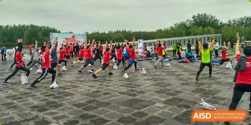 A large group of students in red and black tops participating in an outdoor fitness or exercise activity on a dark paved area, at Ulink College of Beijing.