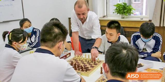 A male teacher with a beard guiding and teaching a group of students how to play chess in a classroom setting, at Ulink College of Beijing.
