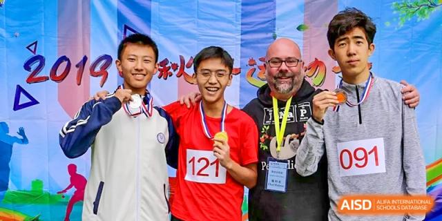 A smiling male student wearing a medal and holding an award next to a bearded adult during a sports or competition ceremony, at Ulink College of Beijing.