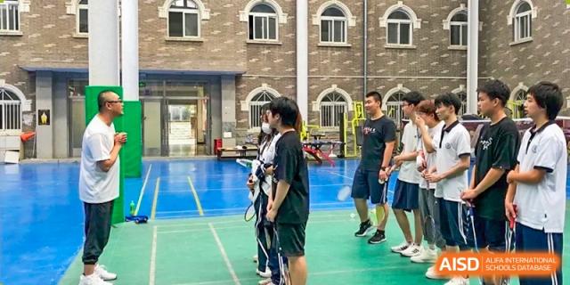 A male student aiming a bow during an outdoor archery practice session on a blue court, with a coach nearby, at Ulink College of Beijing.
