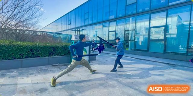 Two students practicing fencing outdoors with foils and protective gear in front of a modern glass building, at Ulink College of Beijing.