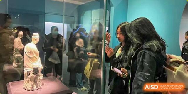 A female student taking a photo or interacting with an exhibit in a brightly lit glass display case indoors, at Ulink College of Beijing.
