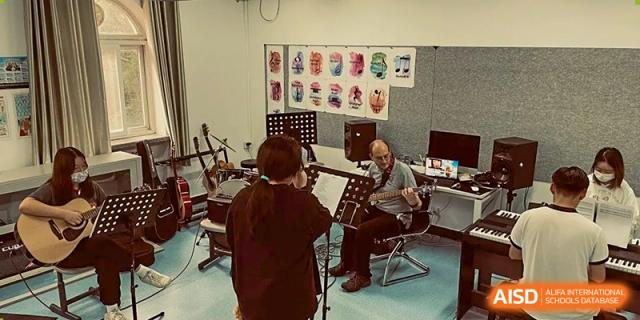 A small group of musicians, including one playing a clarinet, rehearsing together in a music room with colorful masks displayed on the wall, at Ulink College of Beijing.