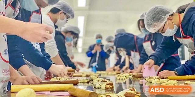 A group of students and instructors in aprons and hats working together at a table, preparing various food items during a cooking class, at Ulink College of Beijing.