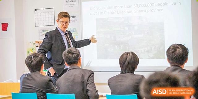 A teacher in a blazer points toward content displayed on a screen for students during a class presentation at Sedbergh School Fuzhou.