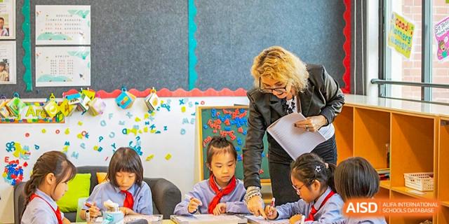 A female teacher assists young primary school students with their work in a brightly decorated classroom at Sedbergh School Fuzhou.
